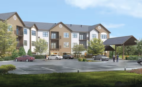 Exterior view of a three-story senior living facility building with beige and white walls, multiple windows, balconies, and a covered entrance. Several cars are parked in the parking lot in front of the building, and a few people are walking near the entrance. Trees and greenery surround the area under a clear blue sky.