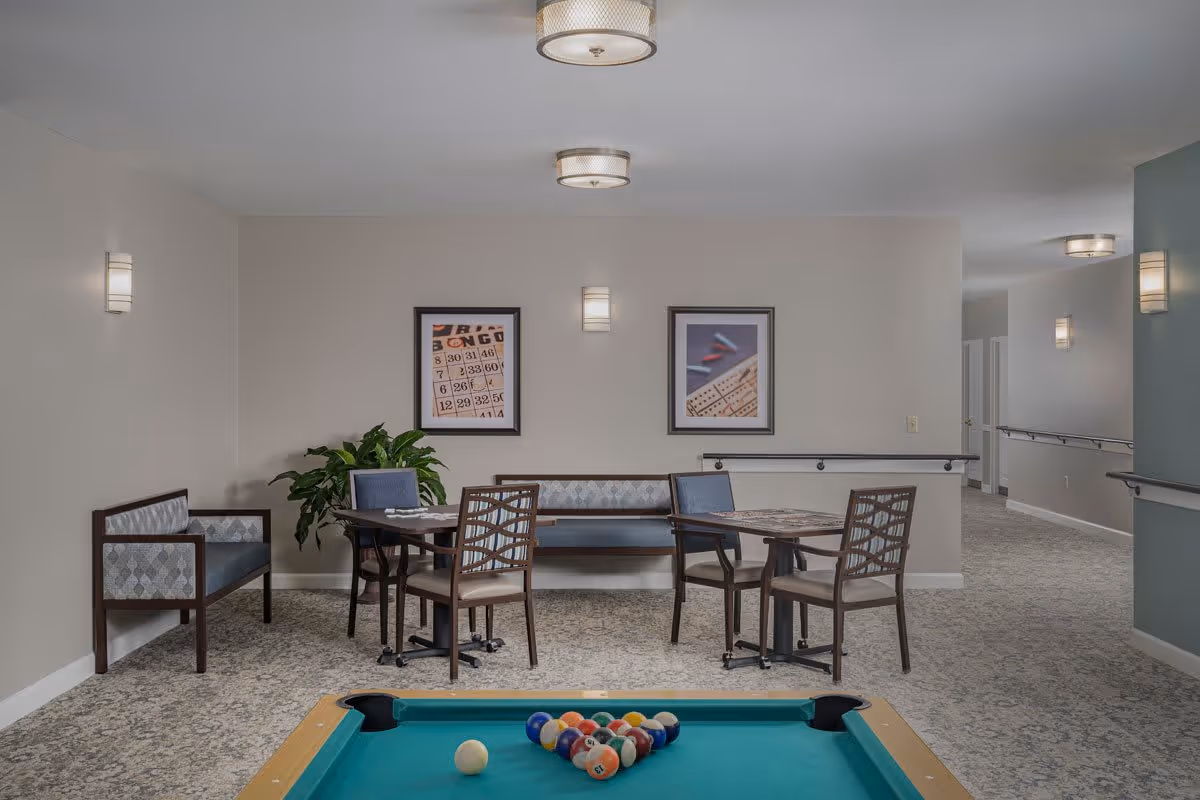 Interior common area with a pool table in the foreground, two small tables with chairs and benches against the wall, framed pictures on the wall, a potted plant, and ceiling and wall lights illuminating the space.