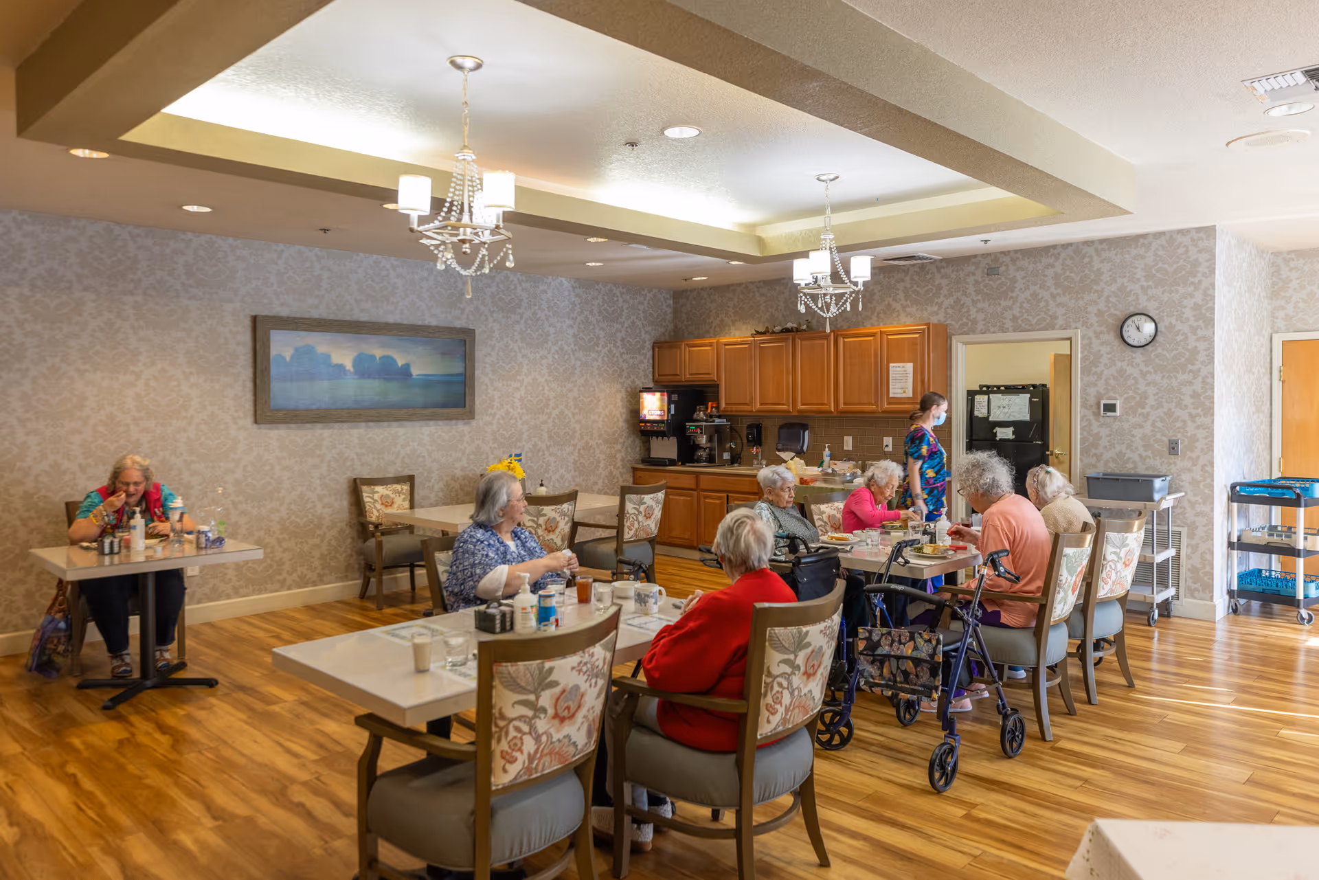 A dining room in a senior living facility with several elderly residents seated at tables eating and drinking. A staff member wearing a mask is standing near the kitchen area with wooden cabinets and a coffee machine. The room has wooden flooring, patterned wallpaper, chandeliers, and a framed landscape painting on the wall.