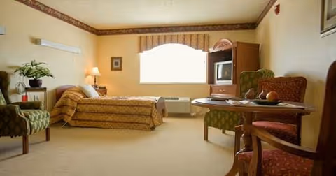 A well-lit bedroom in a senior living facility with a single bed covered in a patterned bedspread, a nightstand with a lamp and flowers, two upholstered chairs, a round table with fruit, and a wooden cabinet with a television. The room has beige walls, a window with a valance, and carpeted flooring.