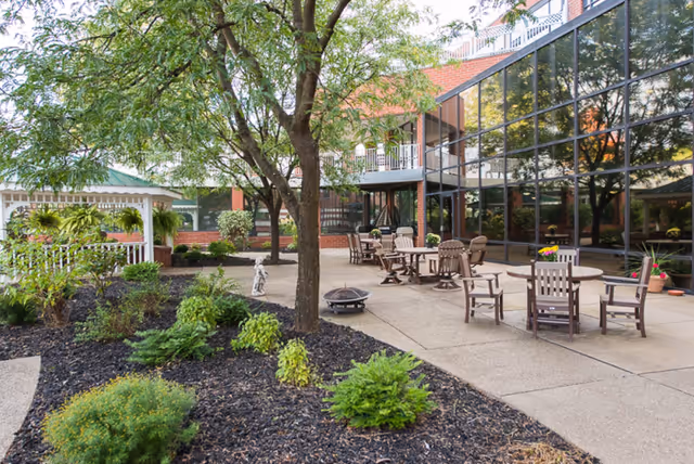 Outdoor patio area with several wooden tables and chairs arranged on a concrete surface. There are trees and landscaped garden beds with green shrubs and mulch. A building with large reflective glass windows is visible in the background, along with a white gazebo on the left side.