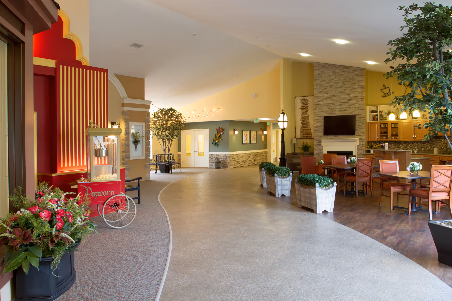 Interior view of a senior living facility common area with a popcorn machine on the left, decorative plants, a curved walkway, and a seating area with tables and chairs near a fireplace and kitchen area on the right.