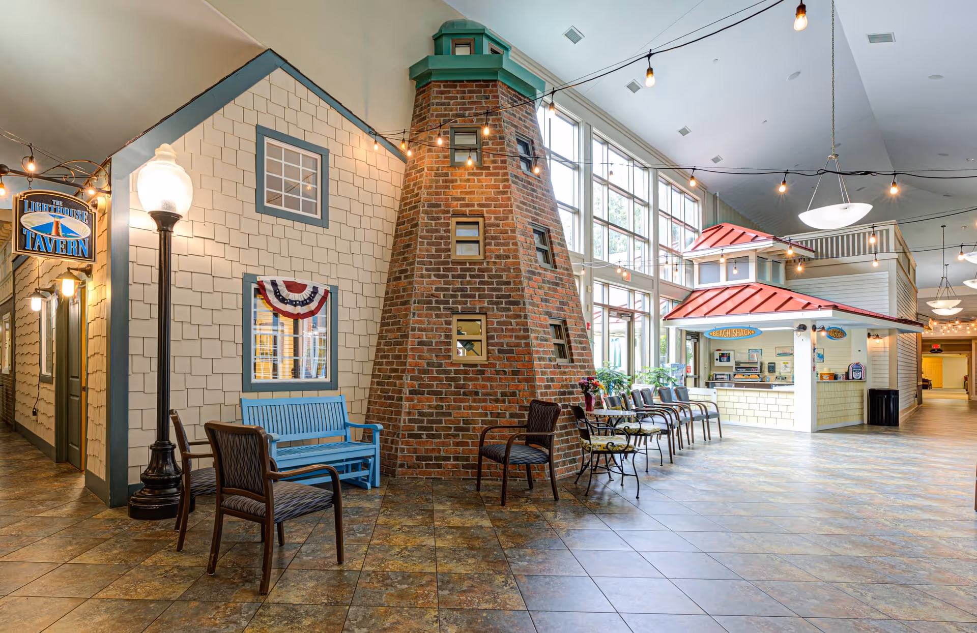 Interior view of a senior living facility featuring a lighthouse-themed brick structure in the center, a blue bench, several chairs, and a small concession stand labeled 'Beach Shack' with large windows letting in natural light.
