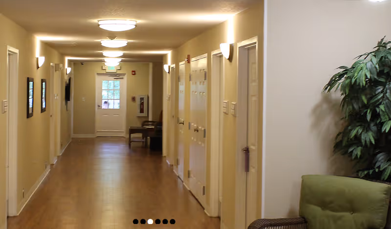 Well-lit hallway in a senior living facility lined with doors, wall sconces, seating at the far end, and a potted plant on the right.