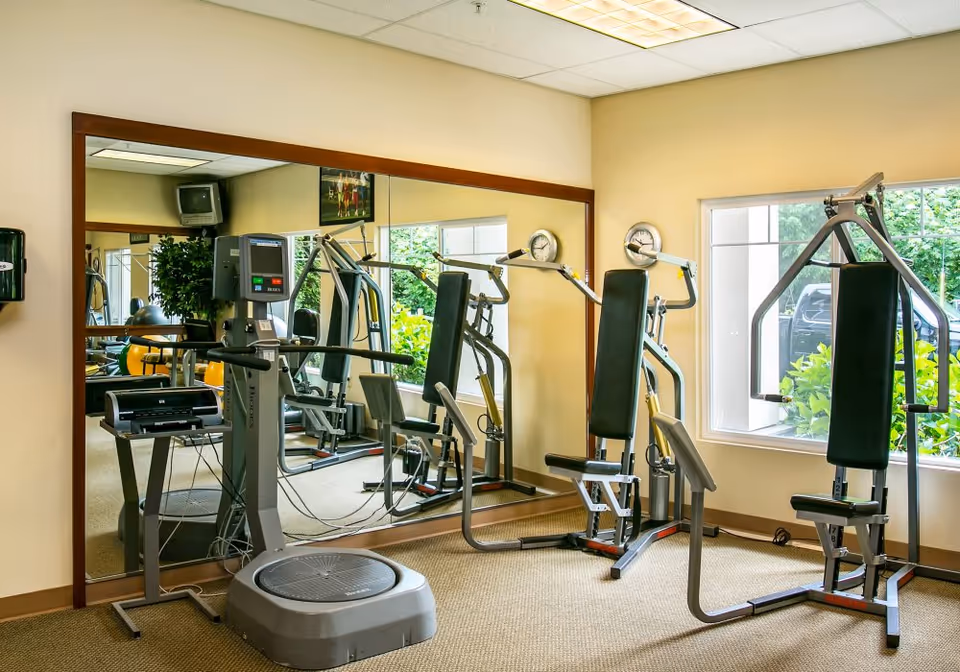 A small fitness room with exercise equipment including a vibration plate machine and a multi-function weight machine. The room has beige walls, a large mirror on one wall, a window showing greenery outside, and a wall-mounted clock.