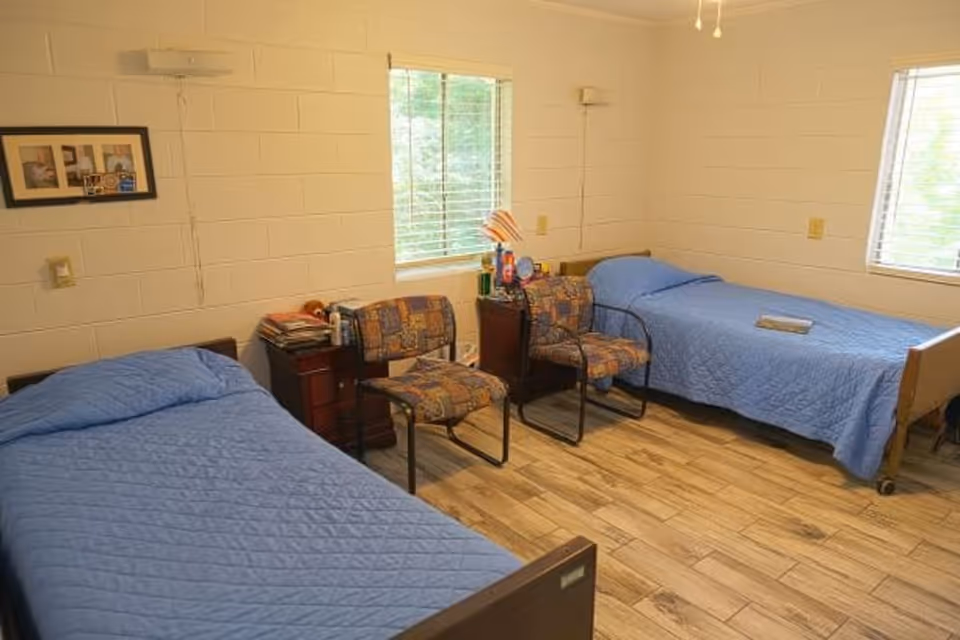 Shared senior bedroom with two twin beds covered in blue quilts, two patterned chairs and a small nightstand between windows.