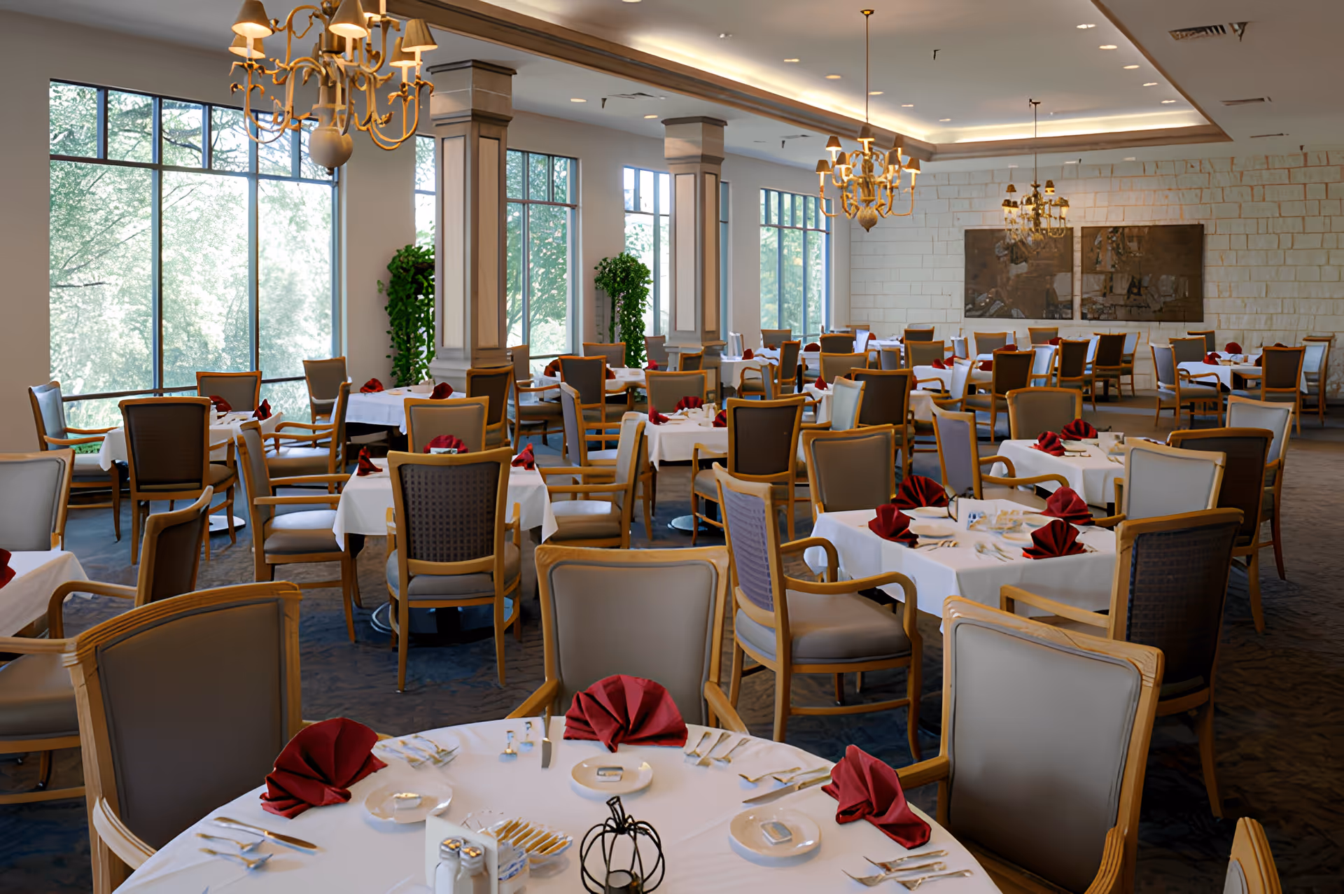 Spacious, elegant dining room with round tables set with white linens and red napkins, large windows, and chandeliers.