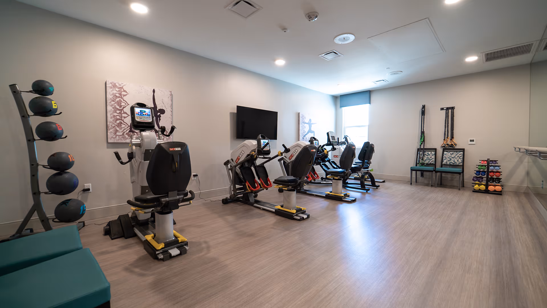 Well-lit fitness room with several recumbent exercise bikes, racks of medicine balls and dumbbells, and seating along the side.
