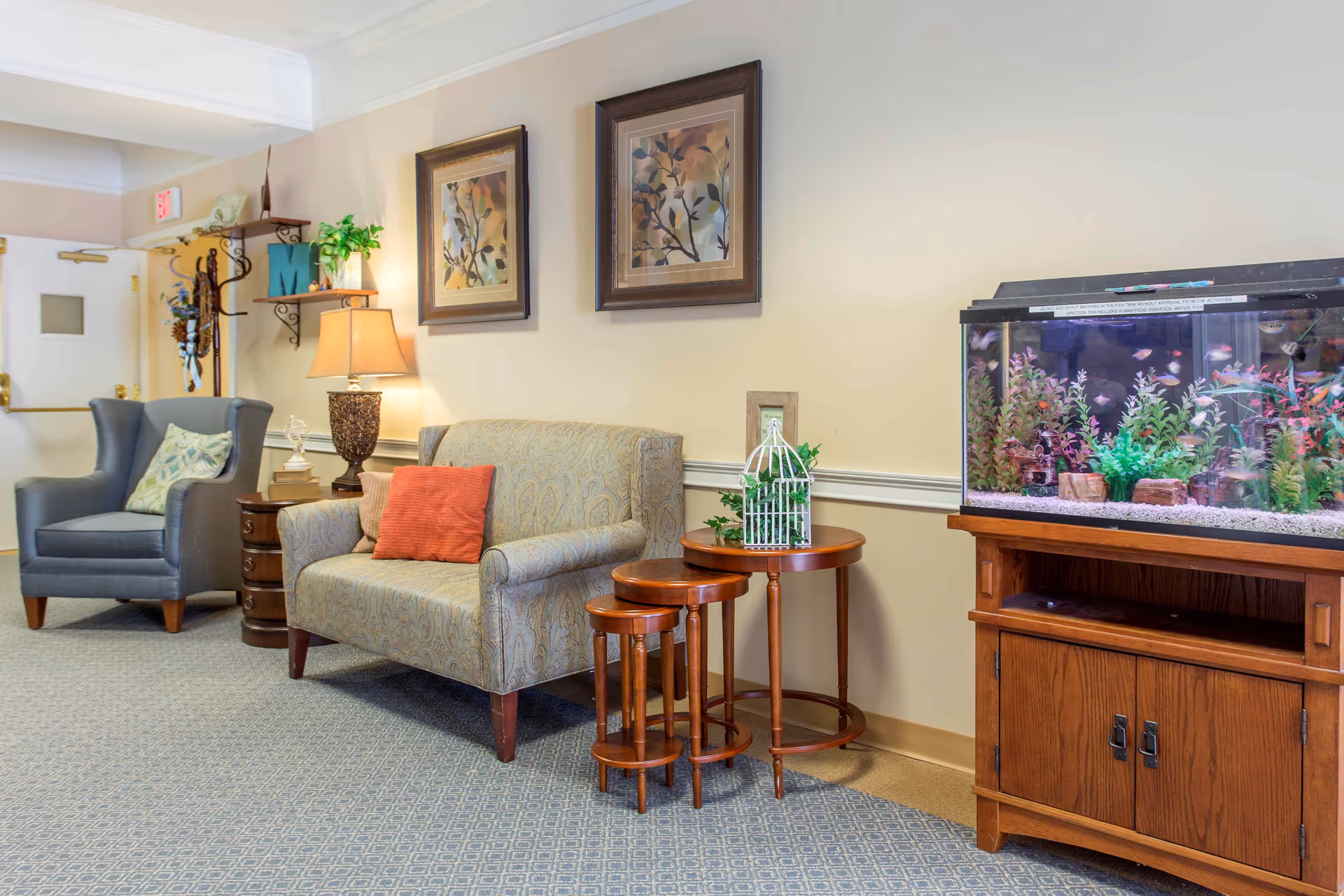 A cozy seating area in a senior living facility with a patterned loveseat, a blue armchair, a set of three nested wooden tables, and a large fish tank on a wooden stand. The walls are decorated with two framed floral paintings, a lamp on a side table, and a coat rack with some hanging items near a door.