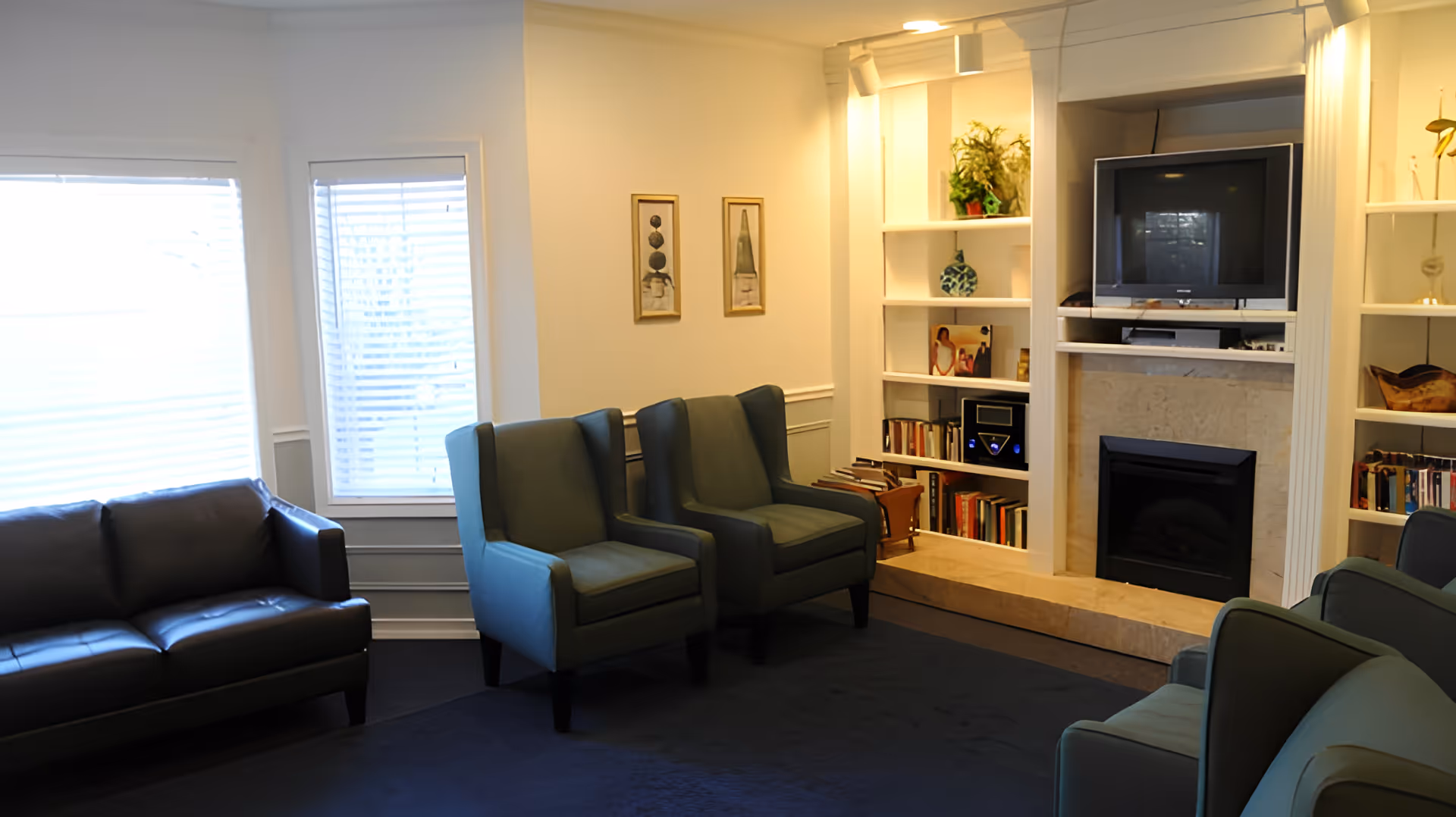 A cozy living room with a dark leather sofa, two green armchairs, a fireplace with a TV above it, built-in white shelves filled with books and decorative items, and two windows with blinds letting in natural light.