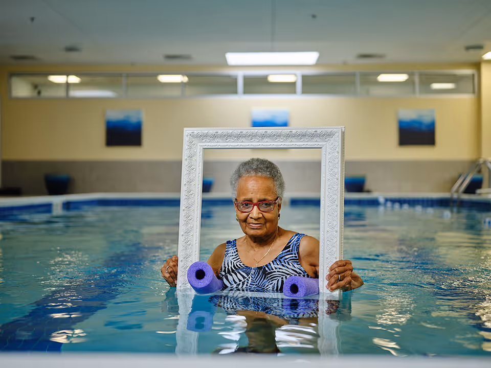 An elderly woman standing in an indoor swimming pool holding a decorative white picture frame in front of her. She is wearing a patterned swimsuit and red glasses, with two purple pool noodles floating in front of her. The background shows the pool area with blue and beige walls and framed artwork.