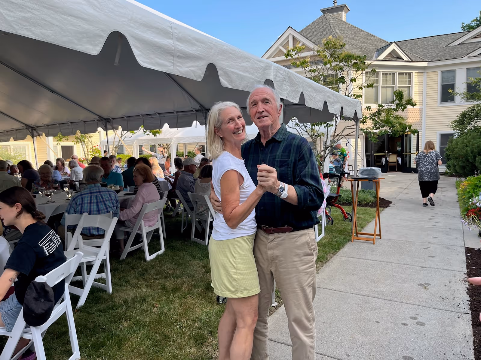 An elderly couple dancing outdoors near a white tent with many people seated at tables under the tent. The setting appears to be a garden or patio area of a senior living facility with a building in the background and a clear blue sky.