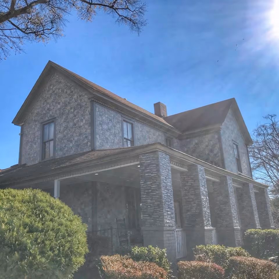 Exterior view of a two-story stone building with a covered porch supported by stone columns, surrounded by bushes and trees under a clear blue sky with sunlight.