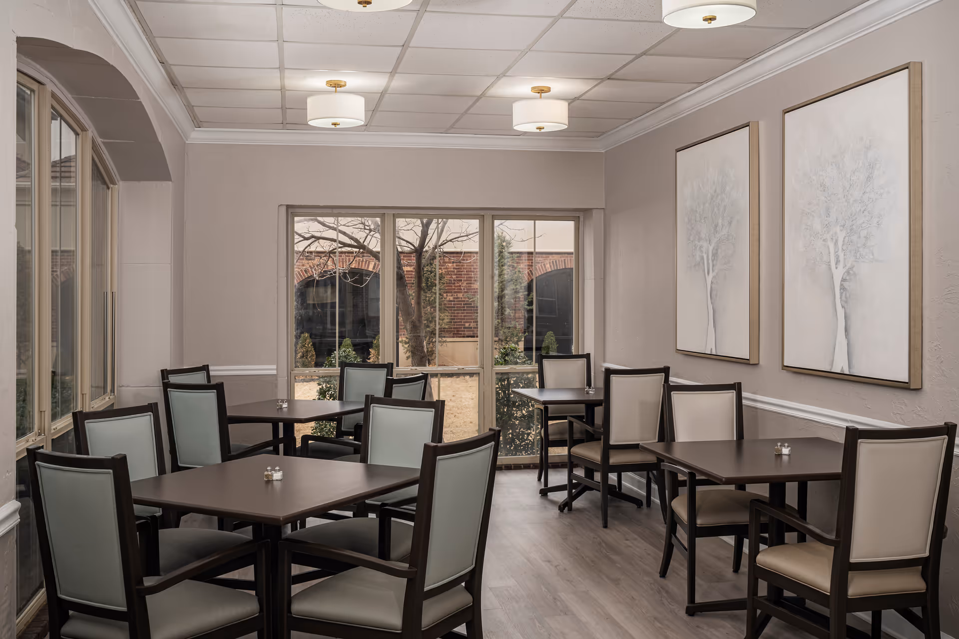 A dining room with several dark wood tables and chairs with light upholstery. The room has large windows showing an outdoor view with trees and brick walls. Two large framed paintings of trees hang on the right wall. The ceiling has recessed lighting fixtures.