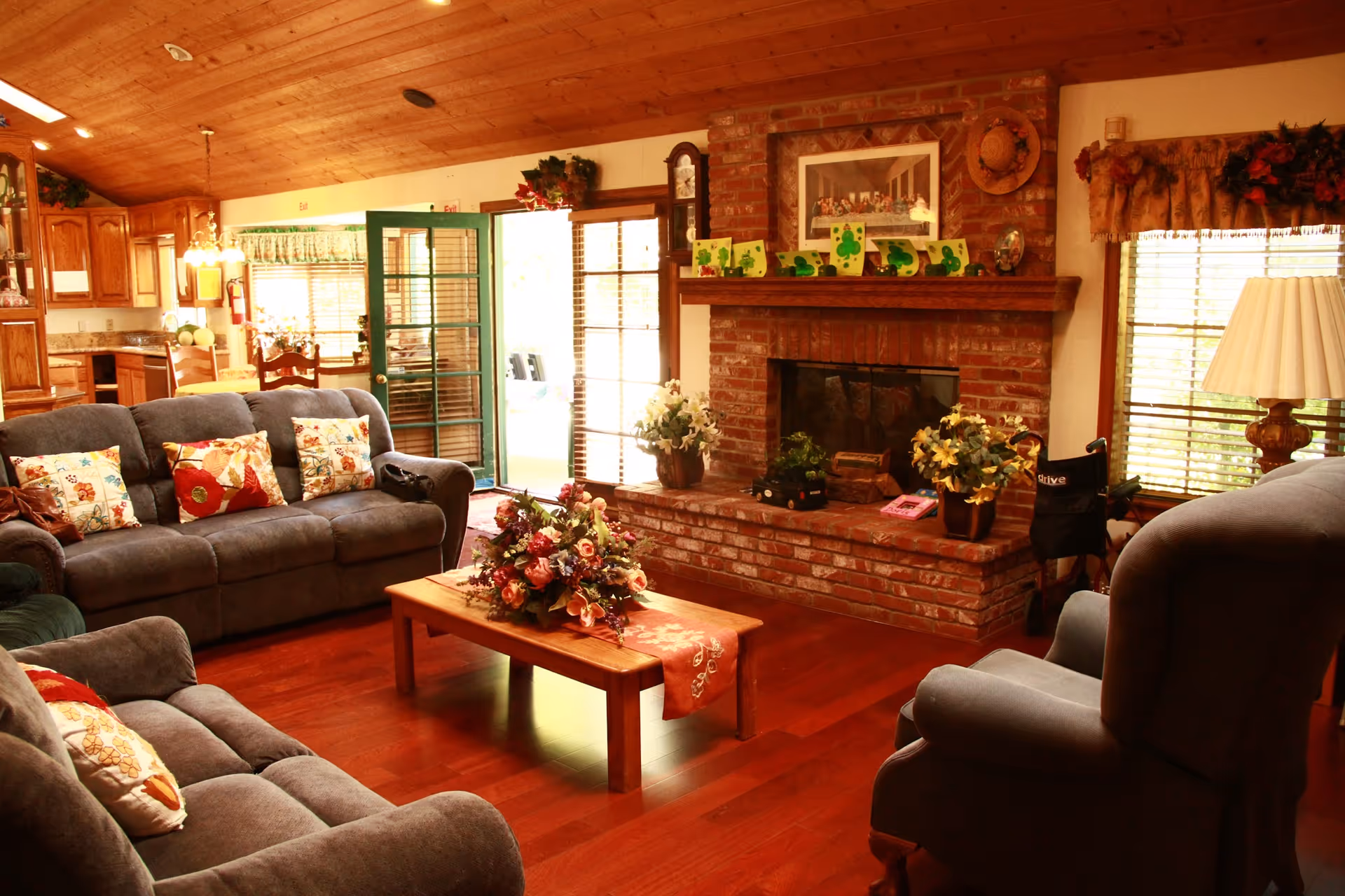 A cozy living room with gray sofas, a wooden coffee table topped with a floral arrangement, and a brick fireplace under a wood-paneled ceiling.