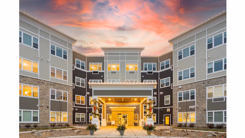 Exterior view of The Preserve of Roseville senior living facility at sunset, showing a modern three-story building with multiple windows lit from inside, a covered entrance with decorative lights, and a paved walkway with potted plants leading to the entrance.