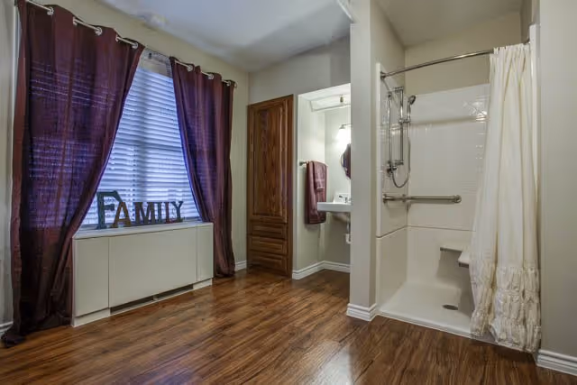 Interior view of a room with wooden flooring, a window with purple curtains, and a white cabinet below the window with decorative letters spelling 'FAMILY'. To the right, there is a small open bathroom area with a shower that has a white curtain and a built-in seat, a sink with a mirror, and a towel hanging on the wall.