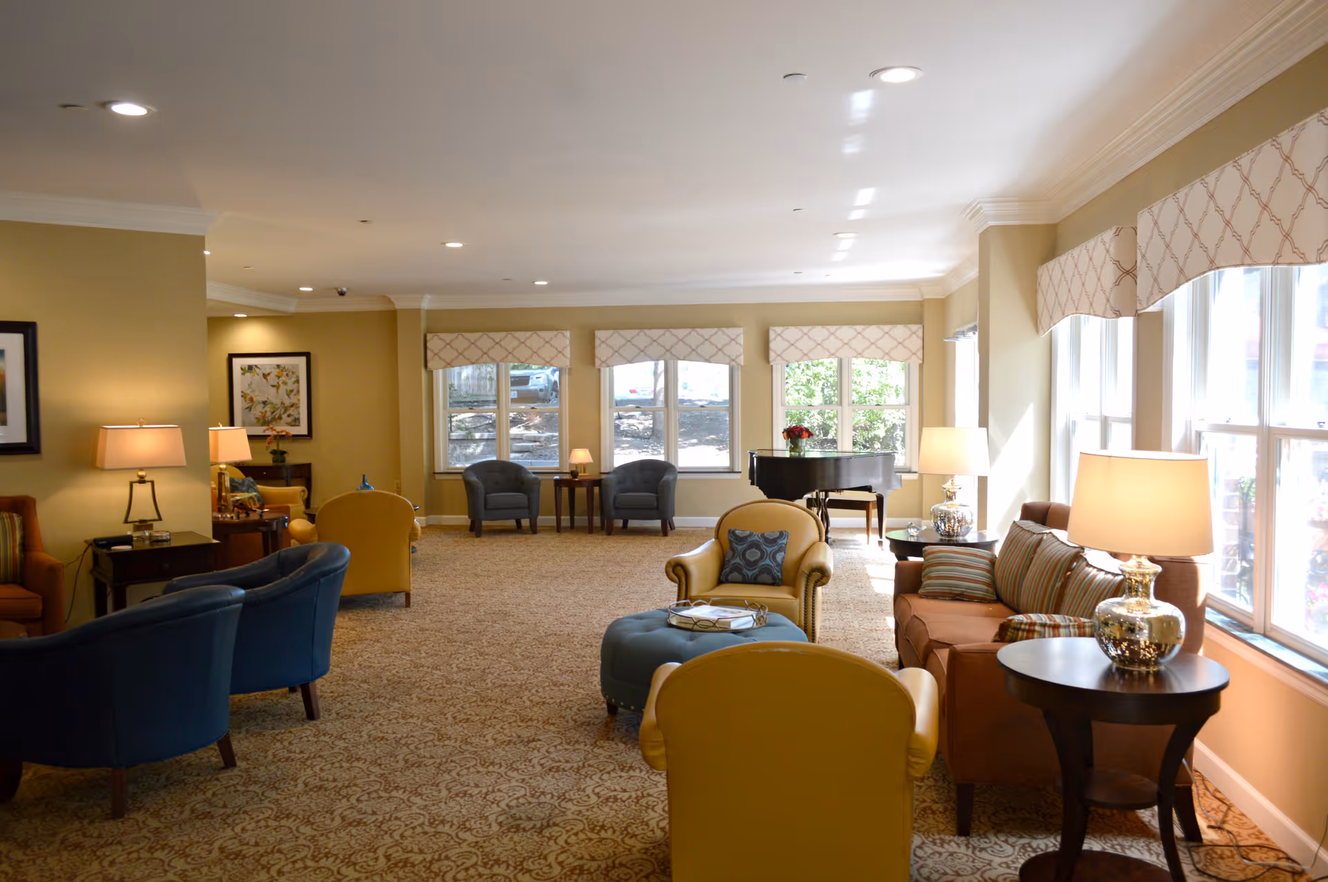 A spacious and well-lit living room area in a senior living facility with multiple armchairs and sofas arranged around small tables with lamps. Large windows with patterned valances allow natural light to fill the room. A grand piano is visible near the windows in the background.
