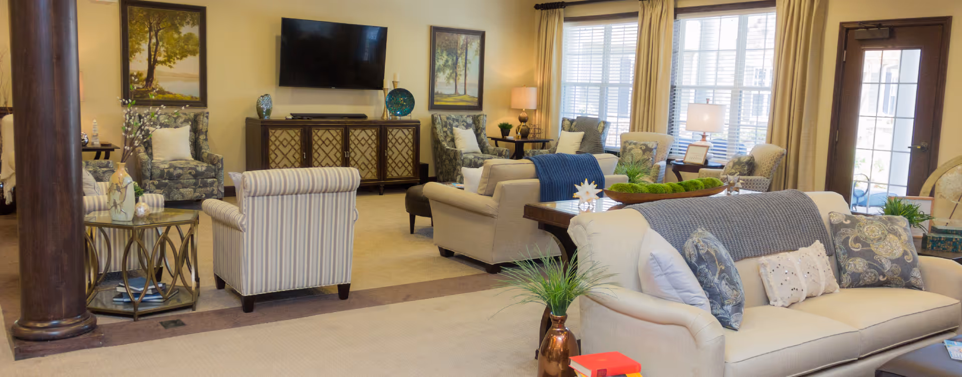 A spacious and well-lit living room area with multiple upholstered sofas and armchairs arranged around a central coffee table. The room features large windows with beige curtains, a flat-screen TV mounted on the wall above a decorative cabinet, framed artwork, and several table lamps providing warm lighting. There are also decorative plants and cushions adding to the cozy atmosphere.