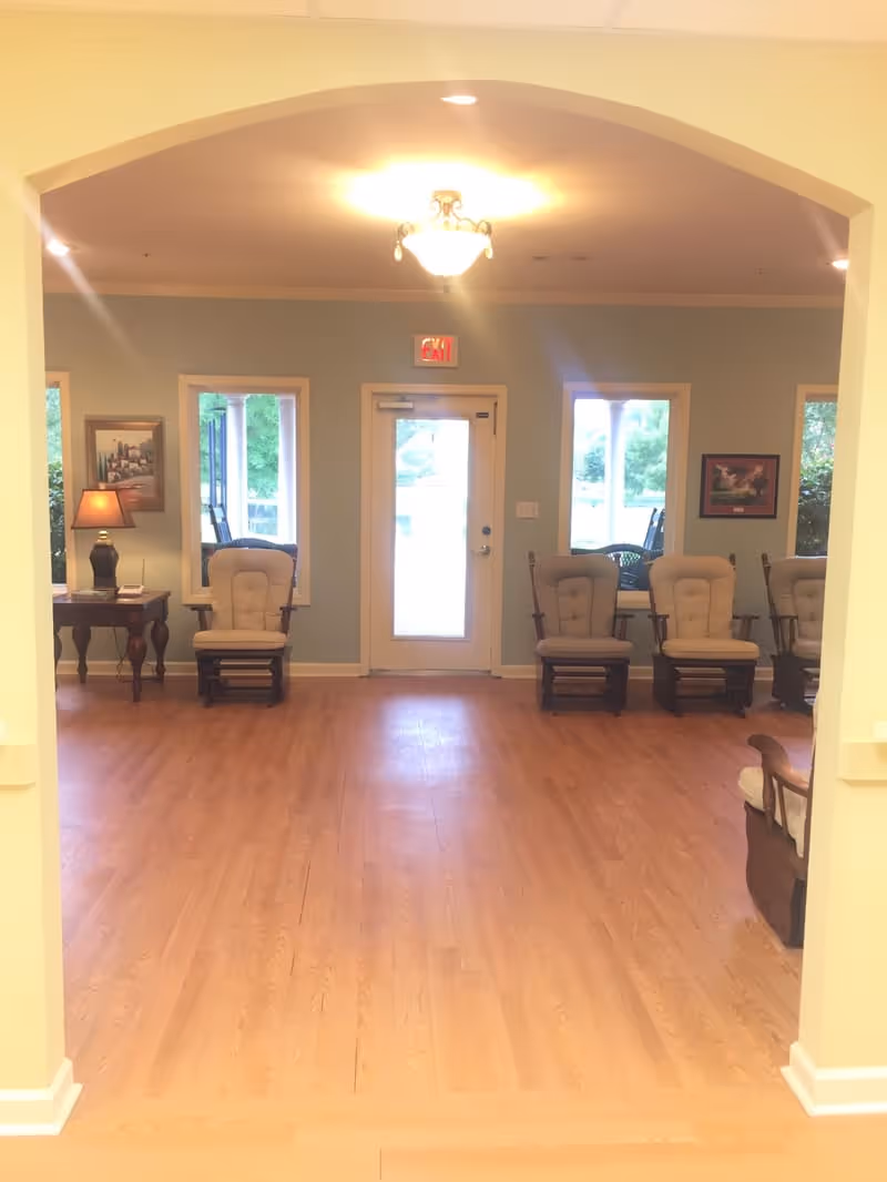 A bright room with light wooden flooring and pale blue walls, featuring a central door with windows on either side. There are several cushioned wooden rocking chairs arranged along the walls, a side table with a lamp and framed pictures hanging on the walls. The ceiling has a decorative light fixture, and the room is viewed through a wide archway.