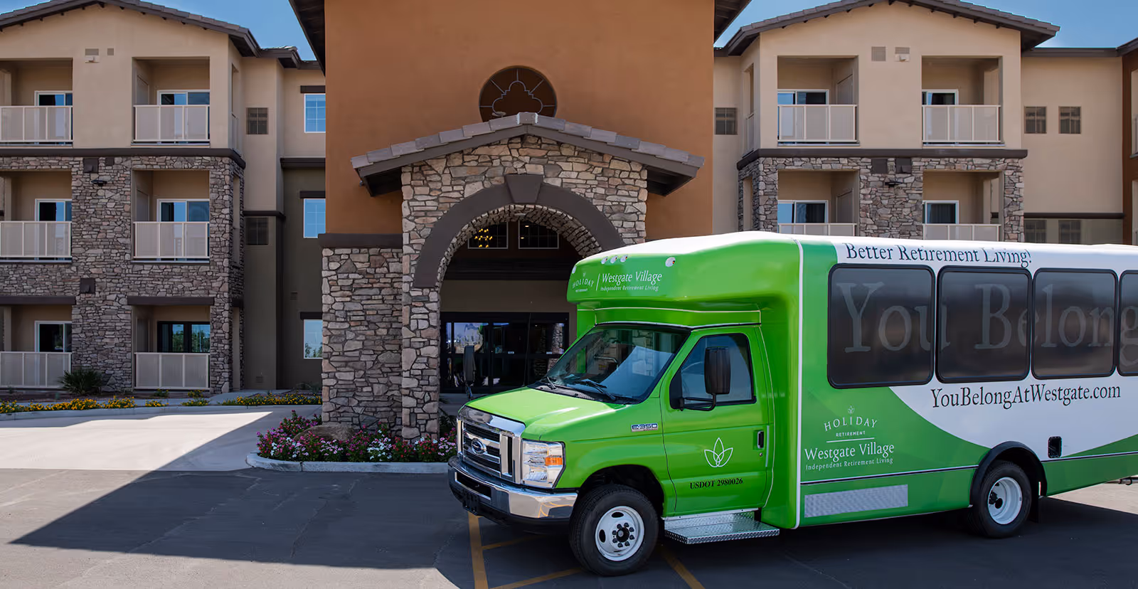 Exterior view of Westgate Village retirement community building with stone and stucco facade and balconies. A green and white shuttle bus with the text 'Better Retirement Living! YouBelongAtWestgate.com' is parked in front of the entrance.