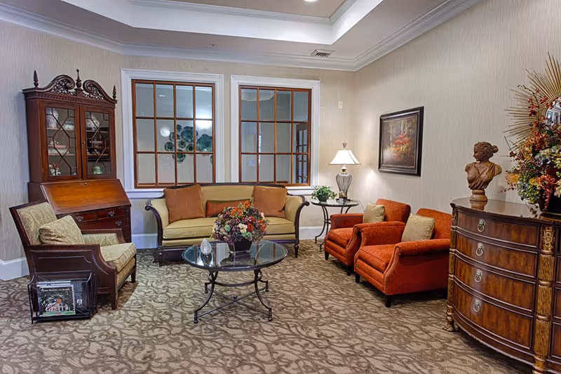A cozy sitting area in an assisted living facility featuring a green upholstered sofa with brown cushions, two orange armchairs with beige pillows, a glass coffee table with a floral arrangement, a wooden cabinet with glass doors, a wooden chest of drawers with a decorative bust and flower arrangement, a side table with a lamp, and two large windows with wooden frames.