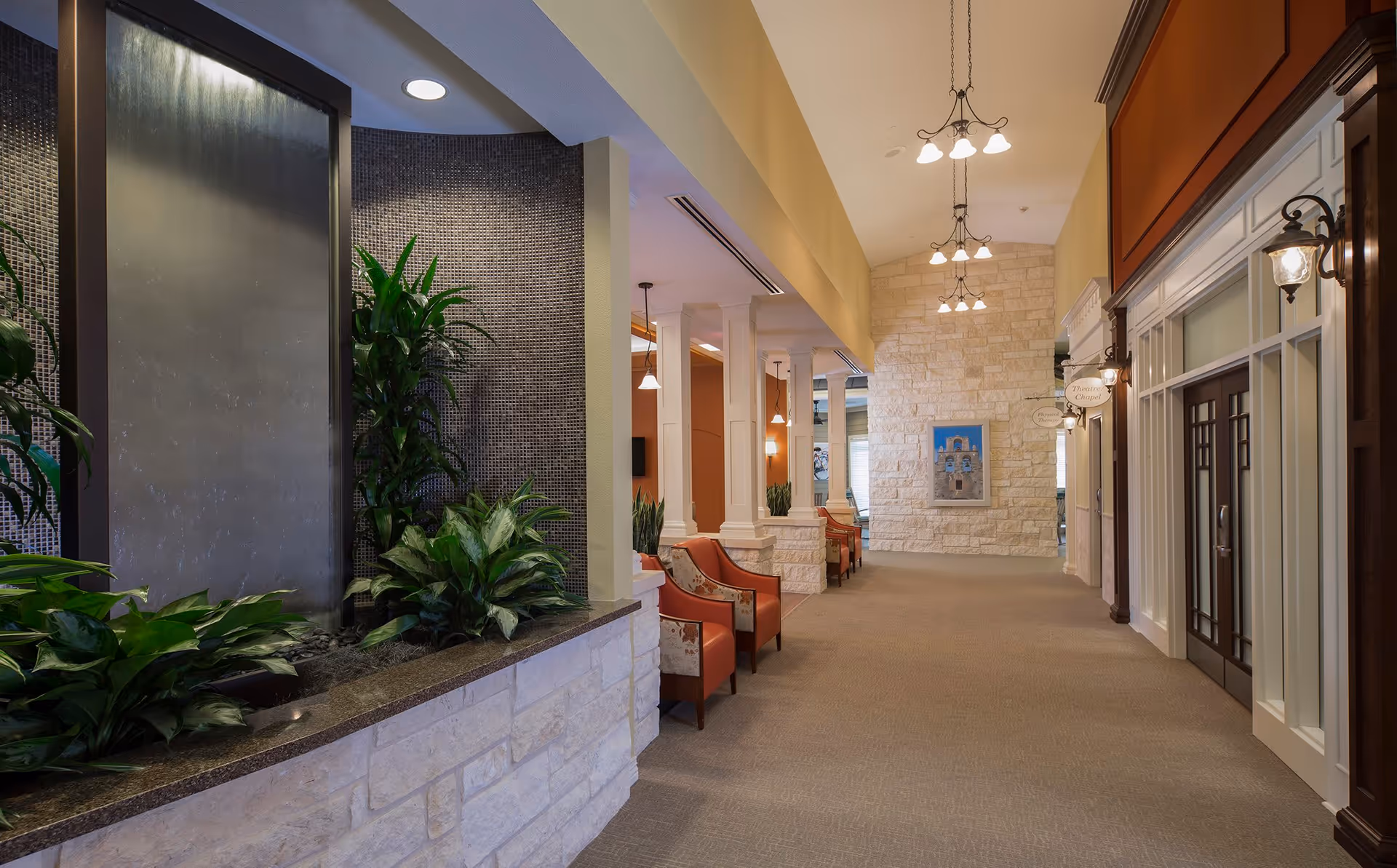 A spacious, well-lit hallway in a senior living facility with a water feature and plants on the left side, comfortable chairs along the wall, decorative hanging lights, and a stone accent wall with artwork at the end.