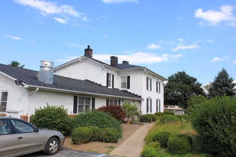 Exterior view of a white, two-story building with a sidewalk, greenery, and a parked car.