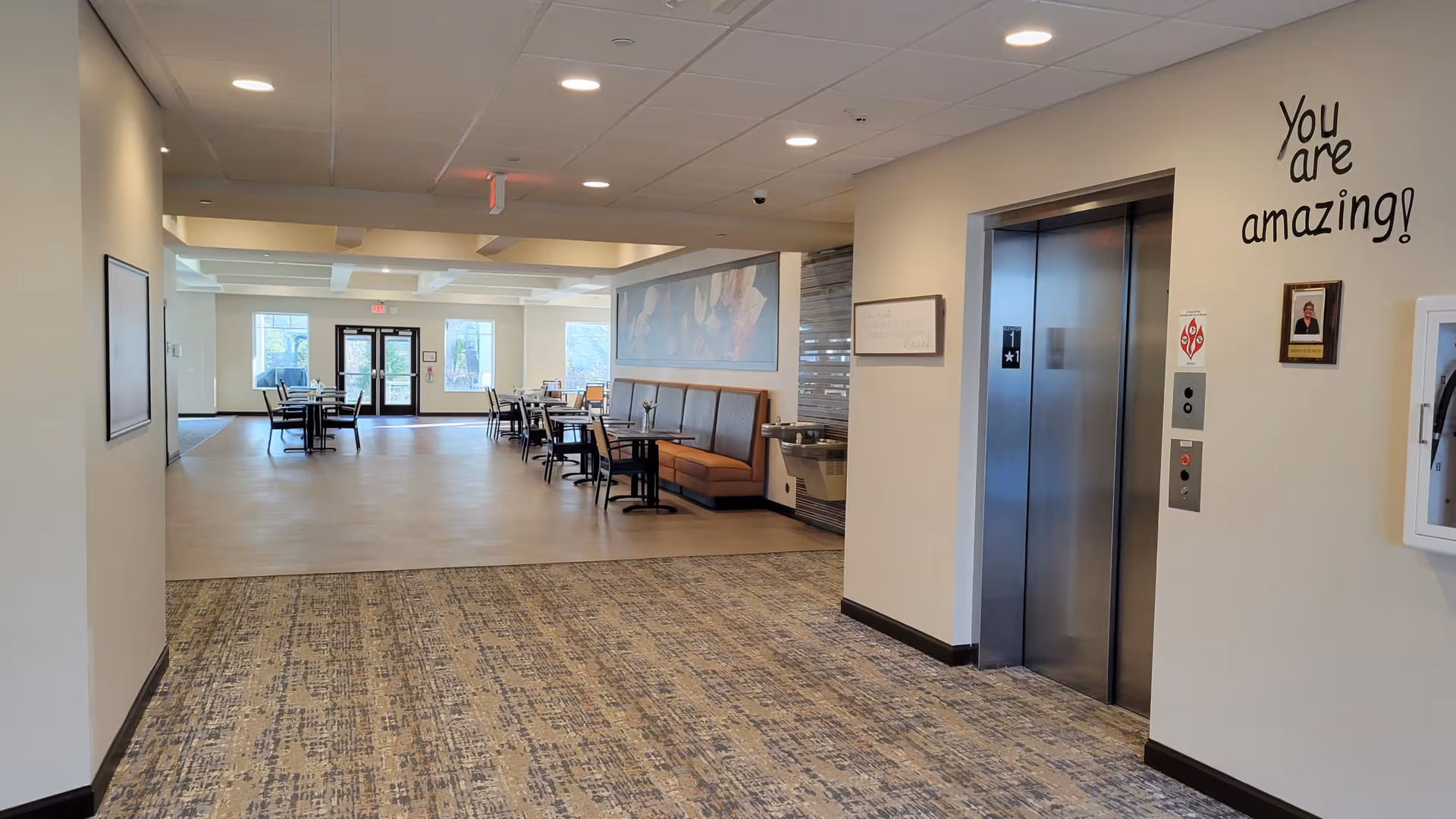 Interior view of a senior living facility hallway leading to a dining area with tables and chairs. On the right side, there is an elevator with a sign above it that says 'You are amazing!'. The floor is carpeted near the elevator and transitions to a smooth surface in the dining area. The space is well-lit with ceiling lights and has large windows at the far end.