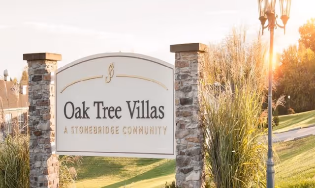 Entrance sign reading "Oak Tree Villas — A Stonebridge Community" set between stone pillars on a landscaped lawn with a lamp post.