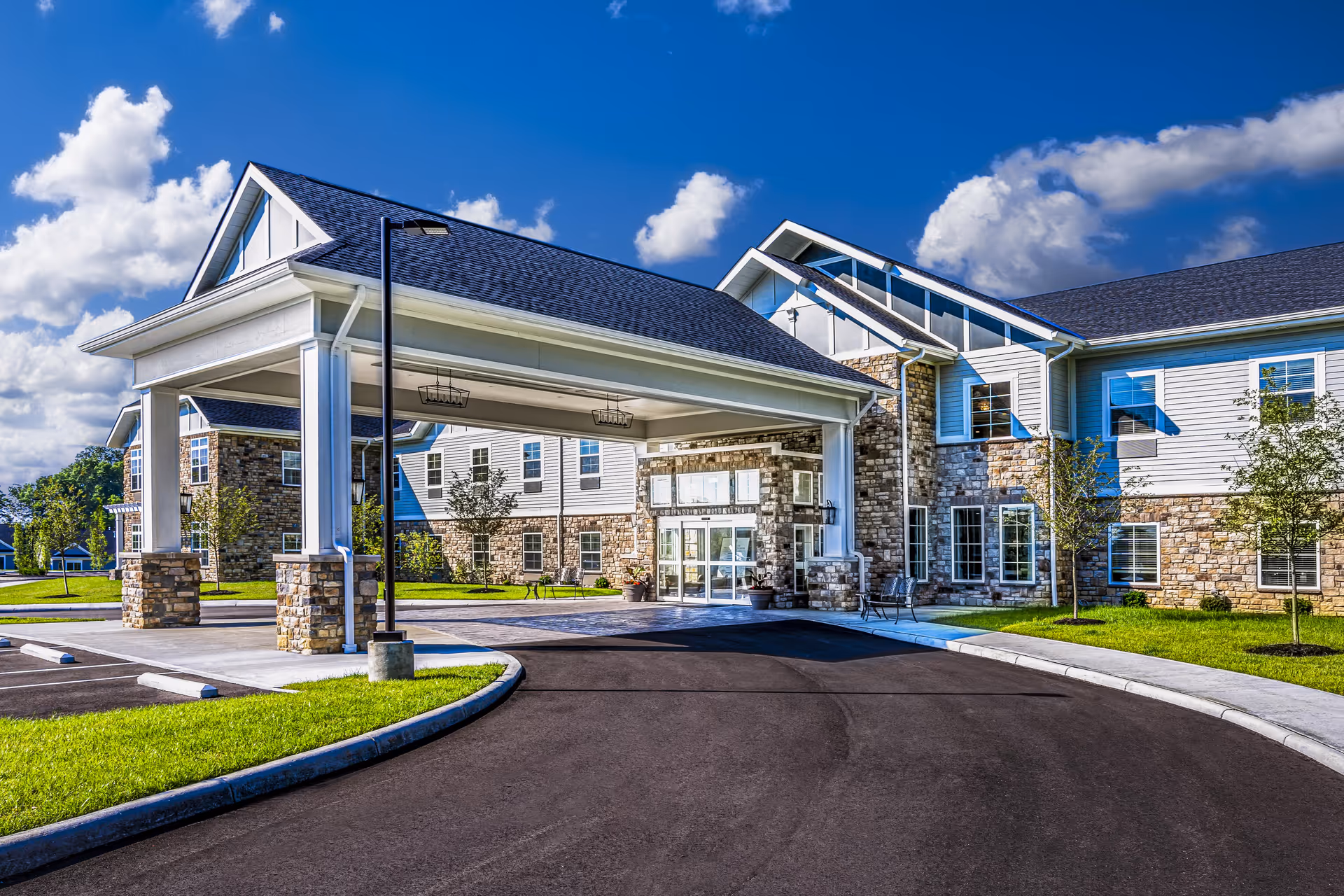 Front entrance of a senior living building with a covered porte-cochère, stone facade, and clear blue sky.