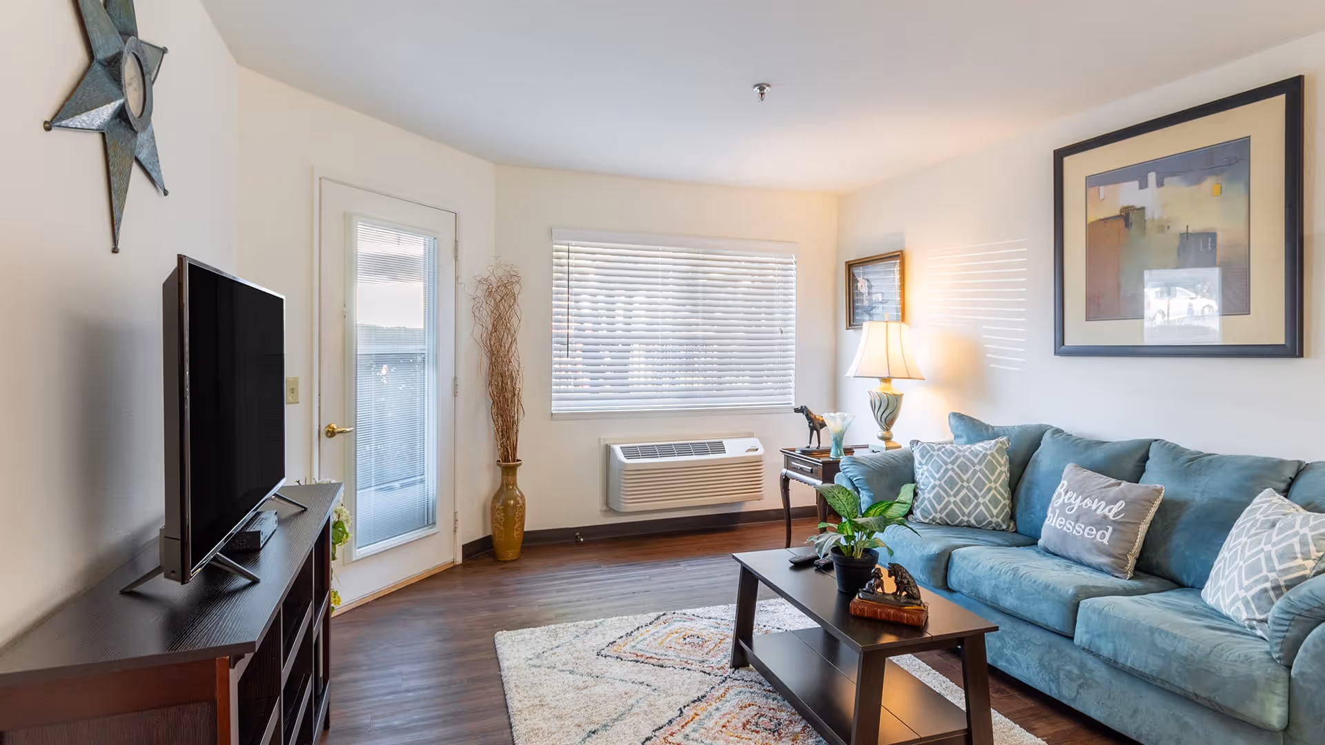 A bright living room with a teal sofa adorned with patterned and text pillows, a dark wooden coffee table with plants and decorative items, a flat-screen TV on a dark wooden stand, a window with blinds, a door with glass panels, a floor vase with dried branches, a table lamp on a side table, and framed artwork on the walls.