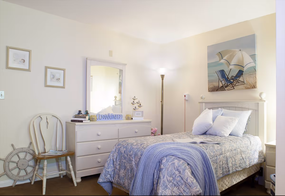 A cozy bedroom with a single bed covered in a blue patterned comforter and a light blue knitted throw. The bed has several white pillows and is positioned against a wall with a painting of two beach chairs under an umbrella. Next to the bed is a white nightstand, and across from the bed is a white dresser with a mirror. On the dresser are decorative items including books and a sign that says 'REMAINED'. A wooden chair and a decorative ship wheel are placed against the wall. A tall floor lamp provides light in the corner of the room.
