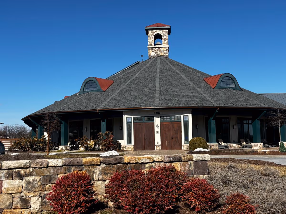 Front view of a single-story octagonal building with a steep shingled roof, small bell tower, stone wall and landscaped shrubs under a clear blue sky.