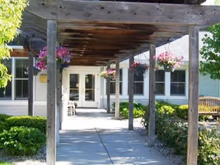 Outdoor walkway with a wooden pergola overhead, hanging flower baskets, and greenery on both sides leading to a building entrance with glass doors and windows.
