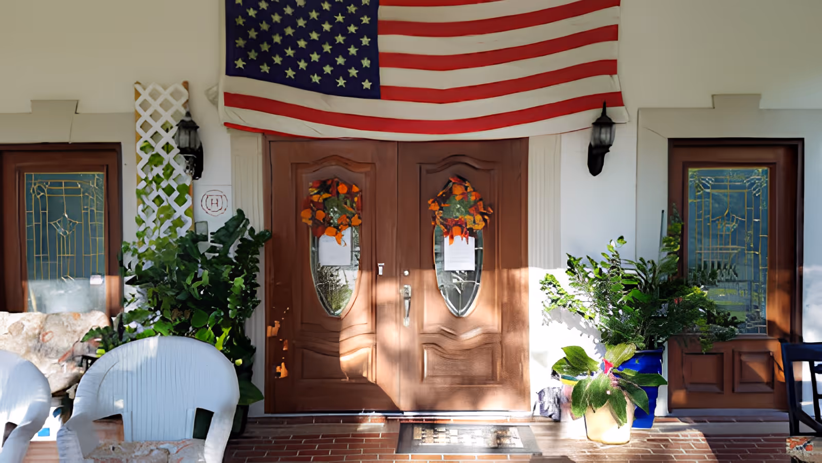 Front porch with double wooden doors decorated with wreaths, an American flag overhead, potted plants and white chairs.
