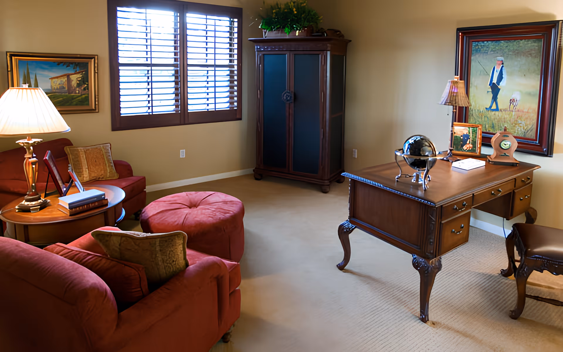 A cozy office or study room with a wooden desk and chair on the right, a red upholstered armchair and ottoman on the left, a wooden cabinet against the wall, and two framed paintings hanging on the beige walls. A table lamp is on a small round side table next to the armchair, and a window with wooden blinds lets in natural light.