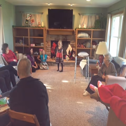 A group of people, including elderly individuals and children, gathered in a cozy living room with a stone fireplace decorated with Christmas stockings. The room has built-in wooden shelves, a large flat-screen TV above the fireplace, and a window letting in natural light. Some people are seated on sofas and chairs, while a young girl stands in the center of the room.