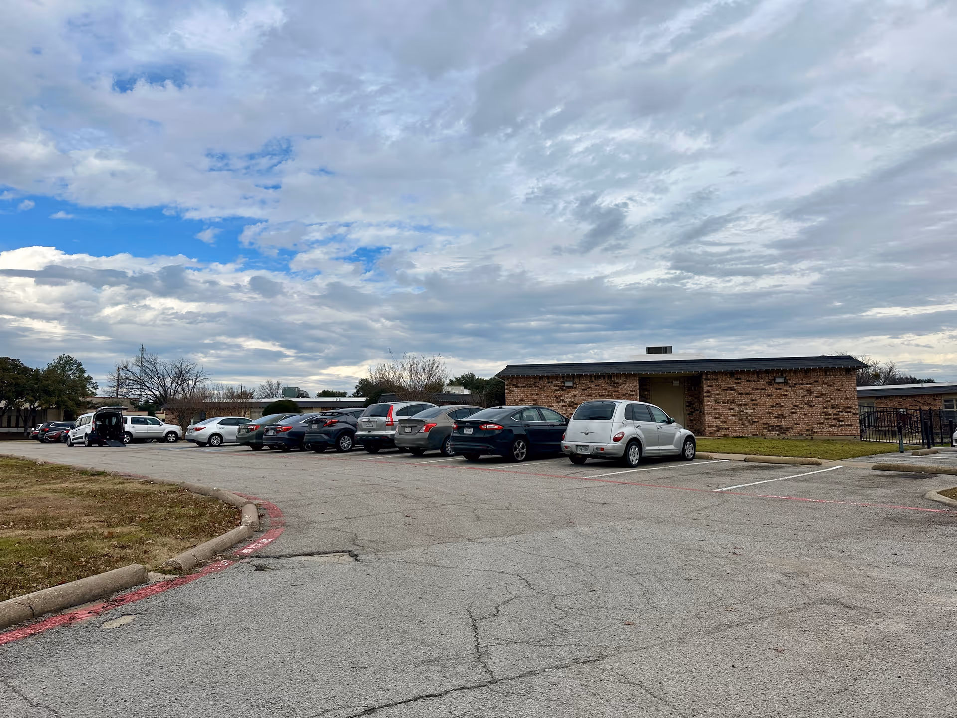 Parking lot with several parked cars in front of a single-story brick building under a cloudy sky.