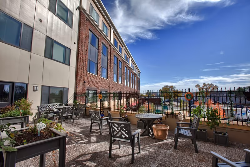Outdoor patio area at a senior living facility with multiple chairs and tables, potted plants, and a black metal fence. The building has a brick and beige exterior, and the sky is partly cloudy with blue patches.