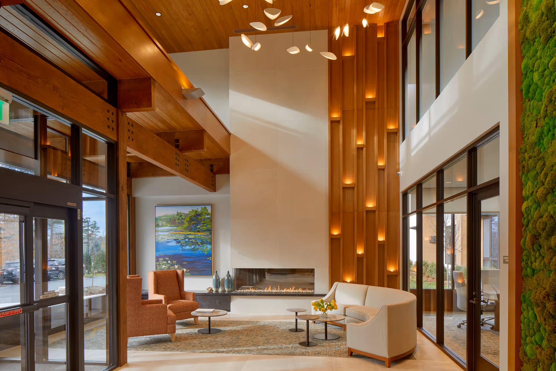 A modern and inviting lobby area with high wooden ceilings and large windows. The space features a contemporary fireplace with a tall white surround, two orange armchairs, a curved beige sofa, and three round coffee tables with a small flower arrangement. There is a large colorful painting on the wall and a green moss wall on the right side. Soft lighting is integrated into the wooden wall panels, creating a warm ambiance.