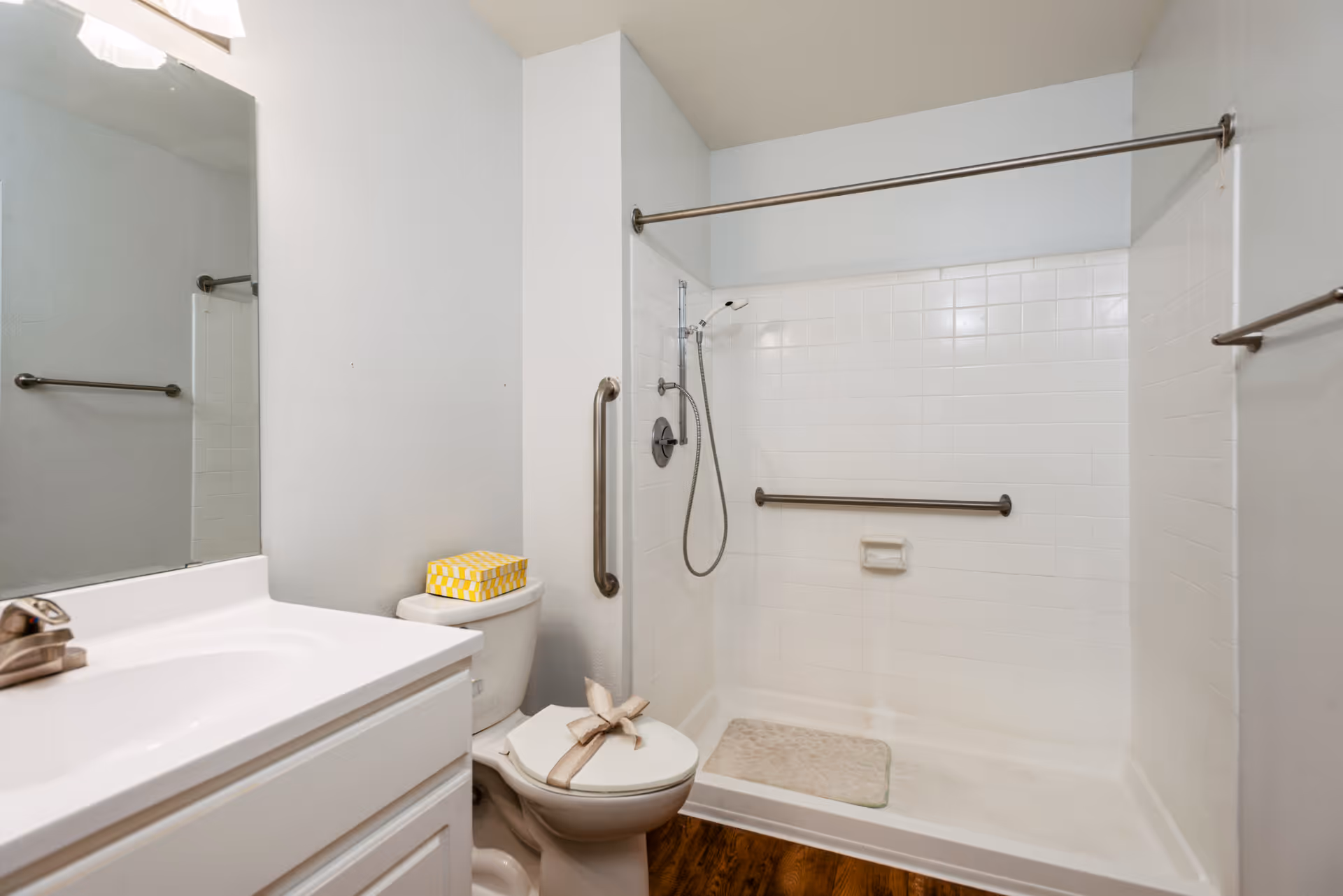 A clean bathroom featuring a white sink with a silver faucet, a toilet with a yellow tissue box on top and a towel folded in a decorative shape on the seat, and a walk-in shower with white tiled walls, a handheld showerhead, grab bars, and a beige bath mat on the floor. The walls are painted light gray and the floor has a wood-like finish.