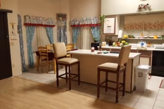 Interior view of a kitchen and dining area in an assisted living home. The kitchen features a central island with two high chairs, a stove, a sink, and various kitchen items on the counters. The dining area has a wooden table with chairs and windows with blue and red curtains. The floor transitions from tile in the kitchen to wood in the adjacent area.