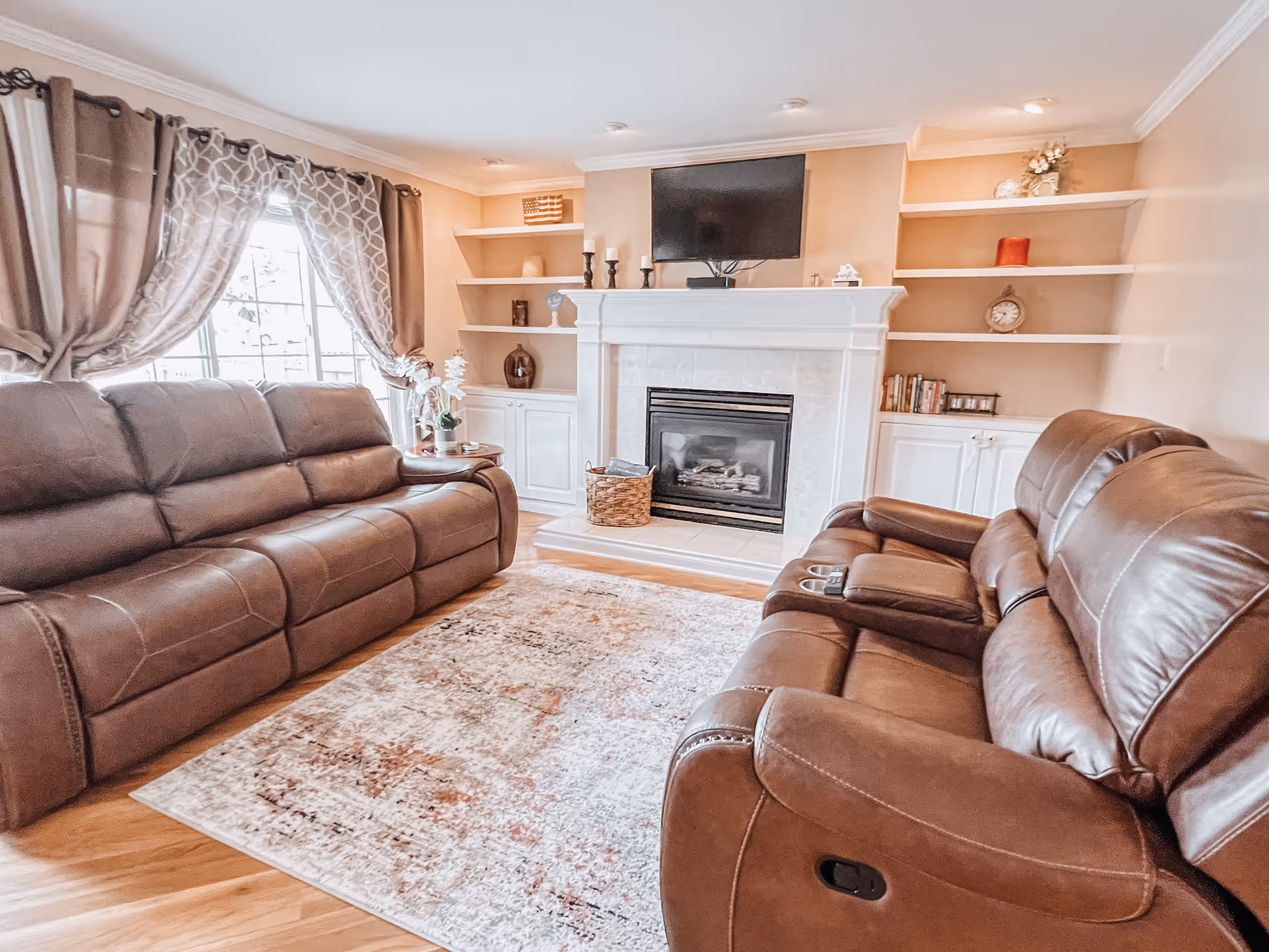 A cozy living room with two brown leather sofas facing each other, a patterned area rug on a wooden floor, a fireplace with a mounted flat-screen TV above it, and built-in white shelves on either side of the fireplace decorated with various items. A window with patterned curtains allows natural light into the room.