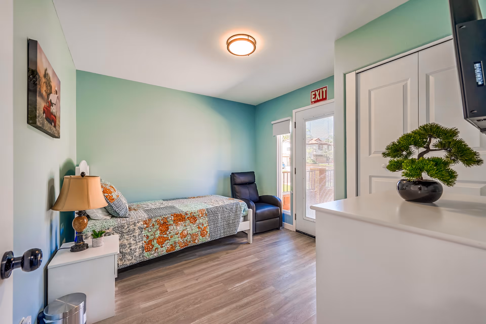 Sunlit single-occupancy bedroom with a bed, armchair, dresser topped with a plant, and an exterior door under an EXIT sign.