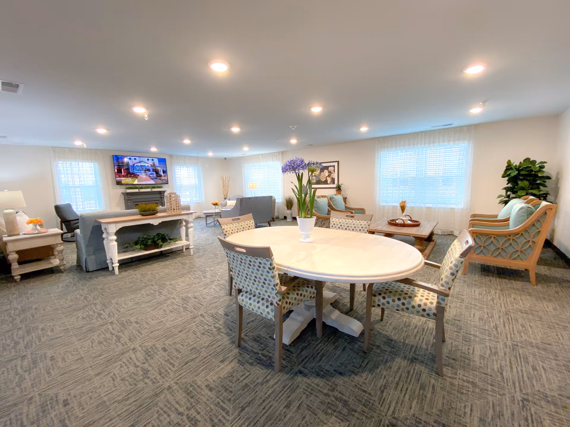 A spacious, well-lit living room area with multiple seating arrangements including sofas and armchairs. There is a round table with four patterned chairs in the foreground, a TV mounted on the wall above a fireplace, and large windows with sheer curtains allowing natural light to fill the room. The room is decorated with plants and framed artwork.