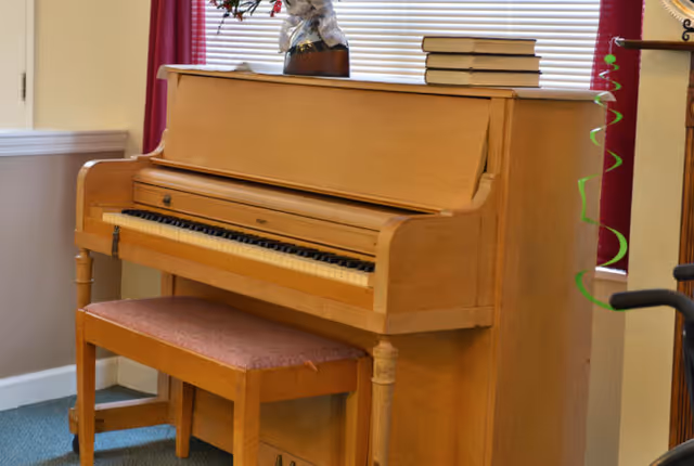 A wooden upright piano with a matching wooden bench in front of it, placed in a room with beige walls, a window with closed blinds, and red curtains. There are some books and a decorative item on top of the piano.