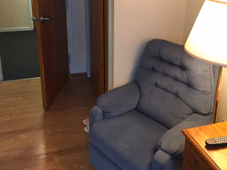 A cozy corner of a room featuring a blue cushioned armchair next to a wooden side table with a lamp and a remote control on it. The floor is wooden, and there is an open door leading to a hallway with carpeted flooring.
