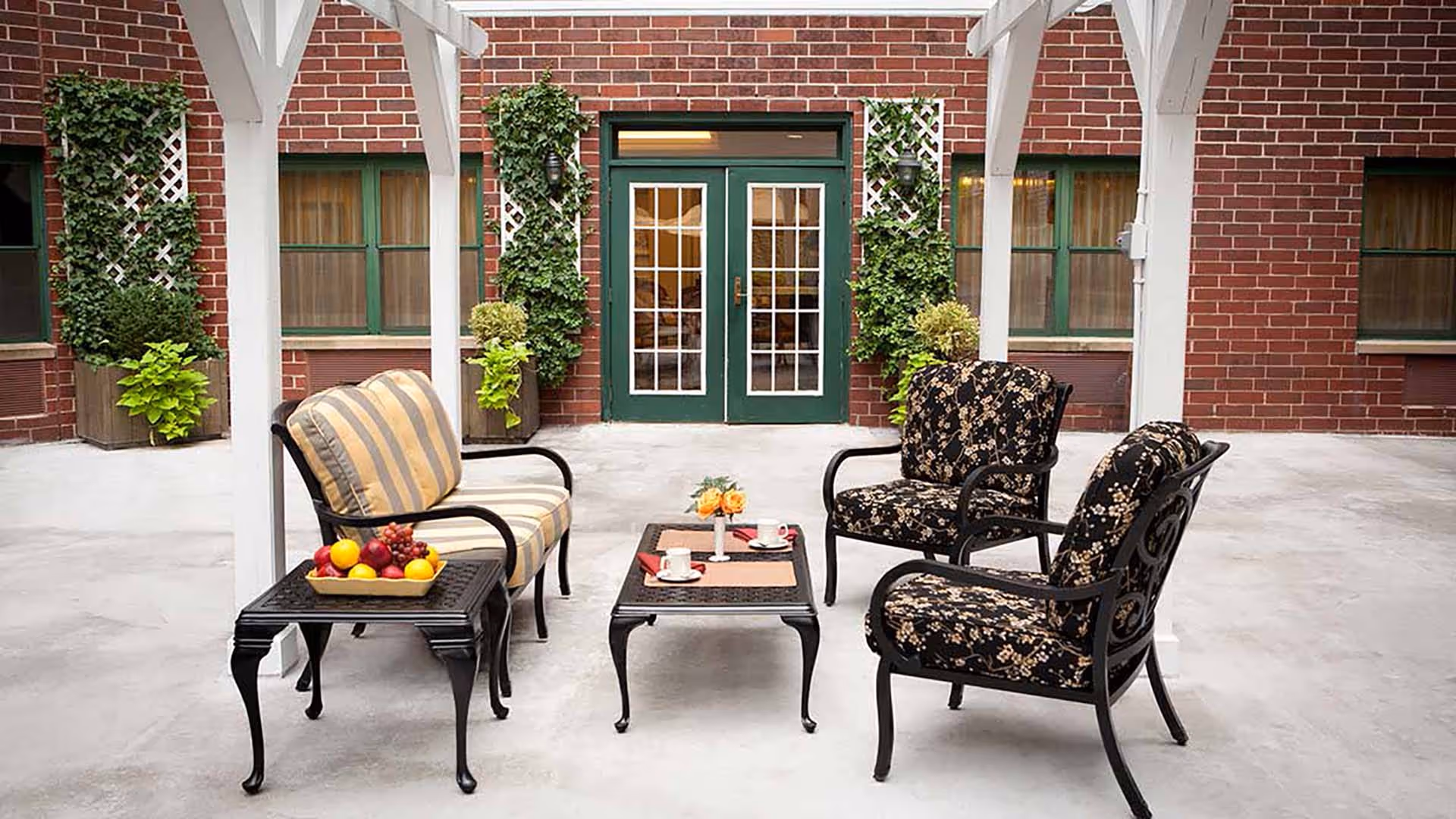 Outdoor courtyard seating area with cushioned chairs and a coffee table in front of a brick building with green double doors.