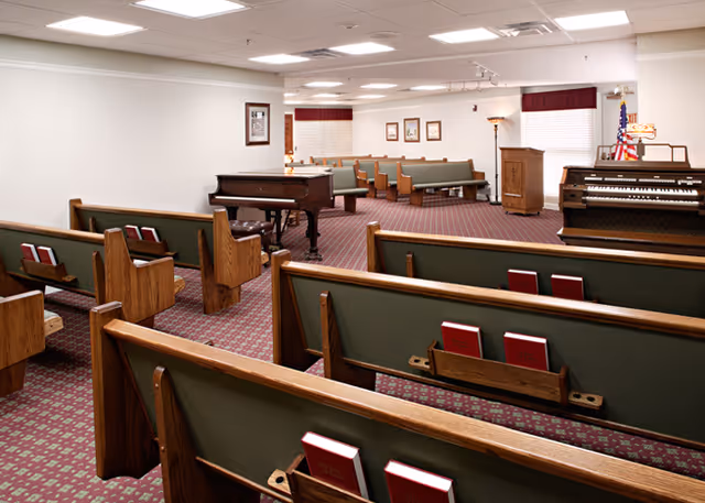 Interior view of a chapel or worship room with wooden pews featuring green cushions and red hymnals, a grand piano, an organ, a wooden podium, framed pictures on the walls, and an American flag in the corner.